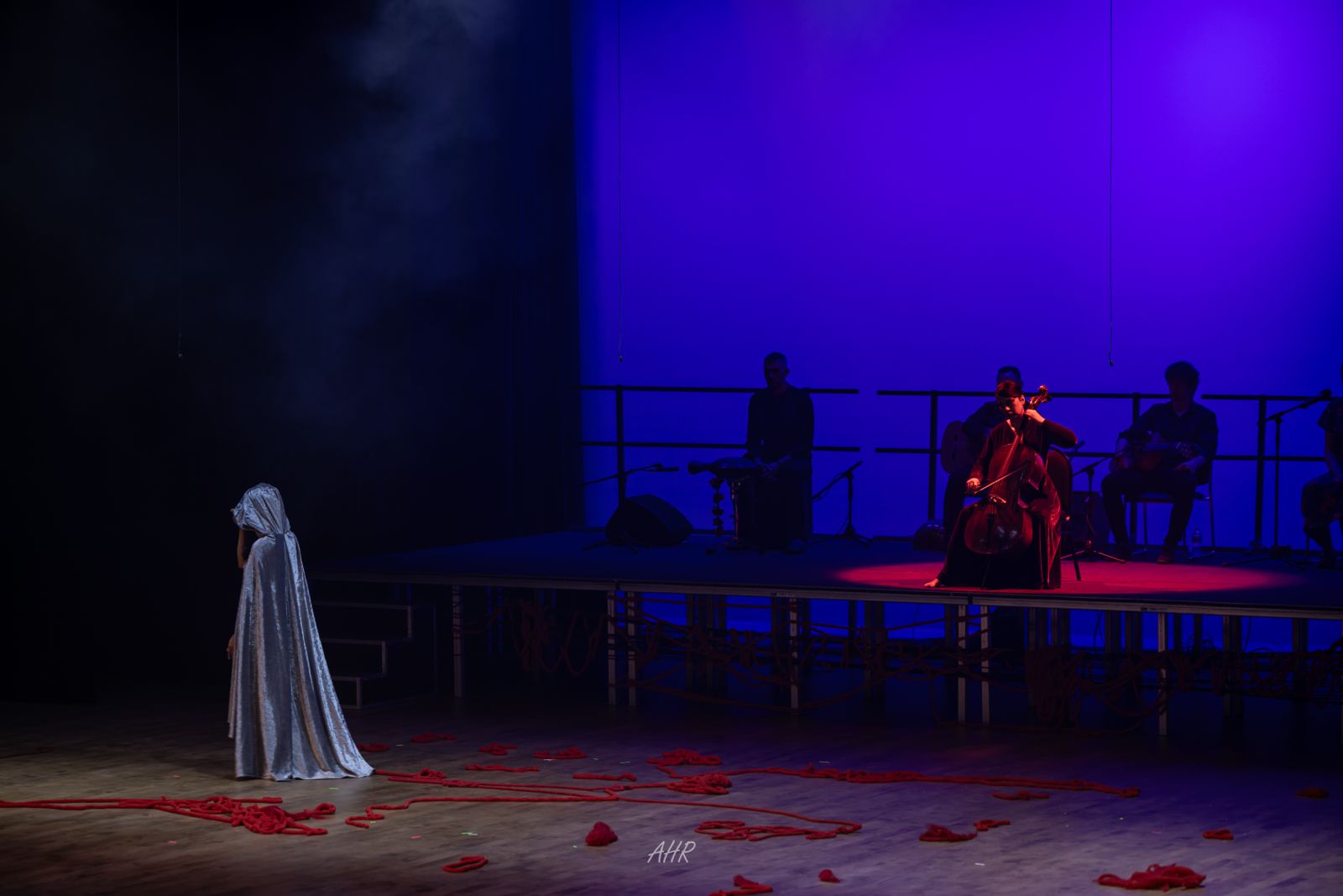 Blood Wedding — Act VI: a cloaked figure at the edge of the stage, red rope strewn across the floor, the cellist lit in red upstage