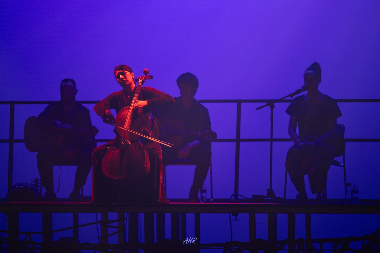 Blood Wedding — Act VI: the musicians in silhouette along the raised platform, the cellist lit red against a deep blue wash