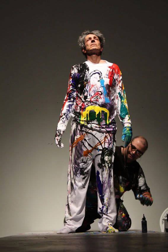 Eternal Braid — a performer standing paint-covered and looking upward, the painter crouching behind him