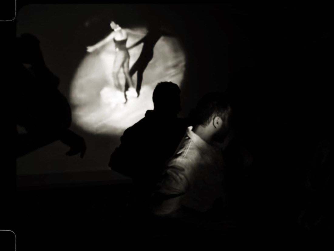Silhouetted figures in a dark room before a projected image of a dancer