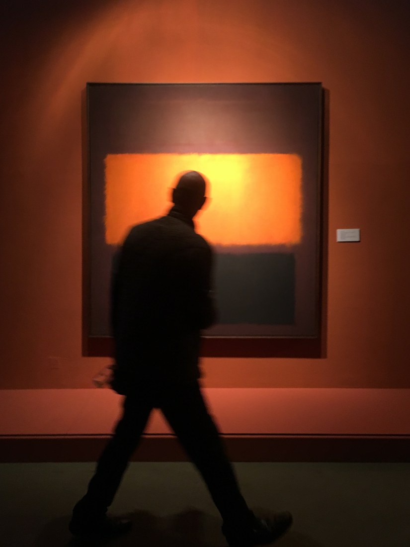 A man walking past a glowing orange colour-field painting in a dark gallery, blurred in motion