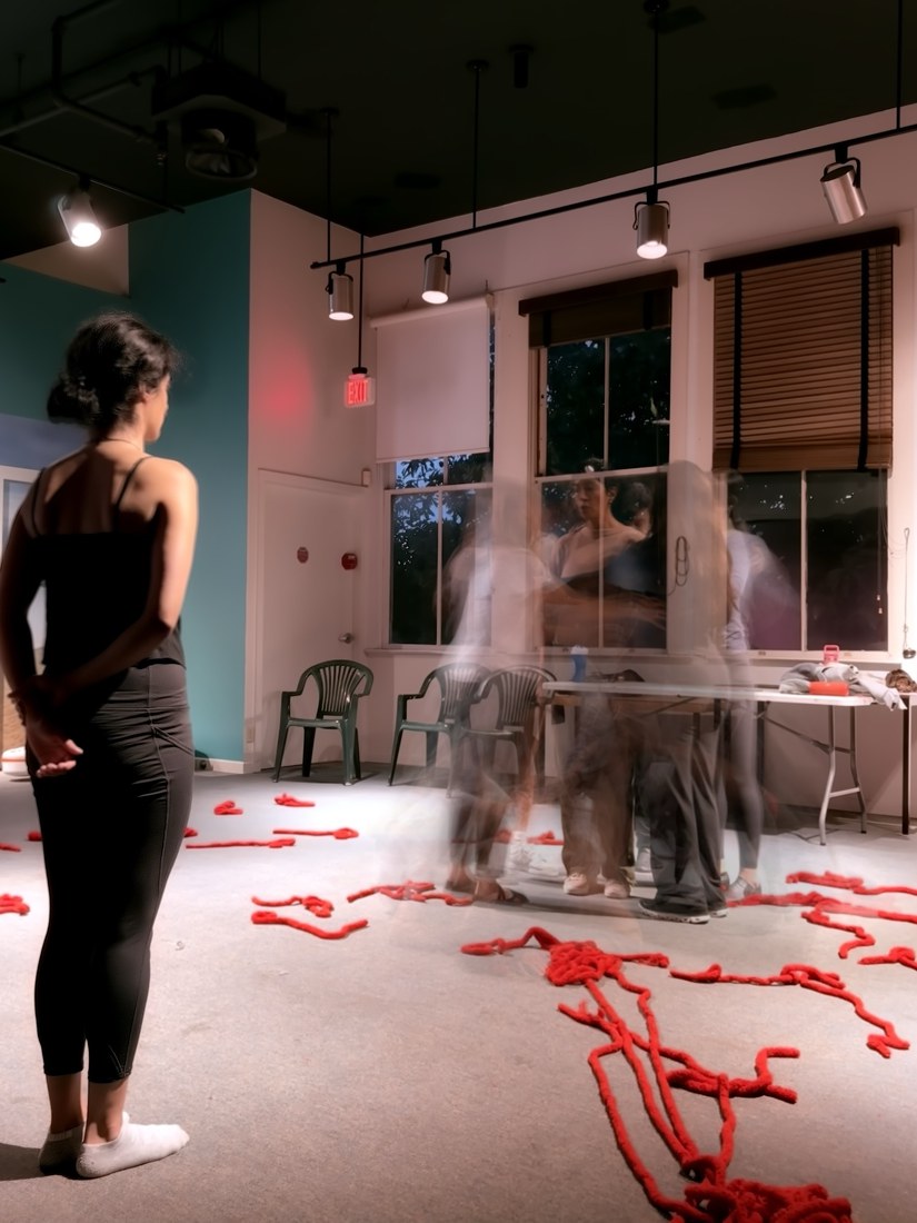 A woman standing still, watching blurred figures move among red rope strewn across a studio floor